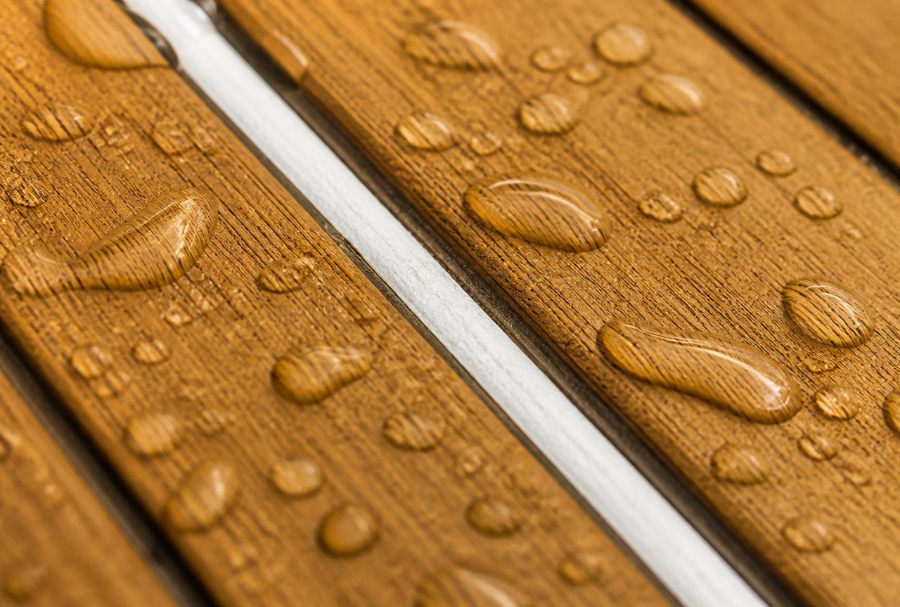 Macro detail of synthetic teak grain with a white caulk seam and beaded water droplets